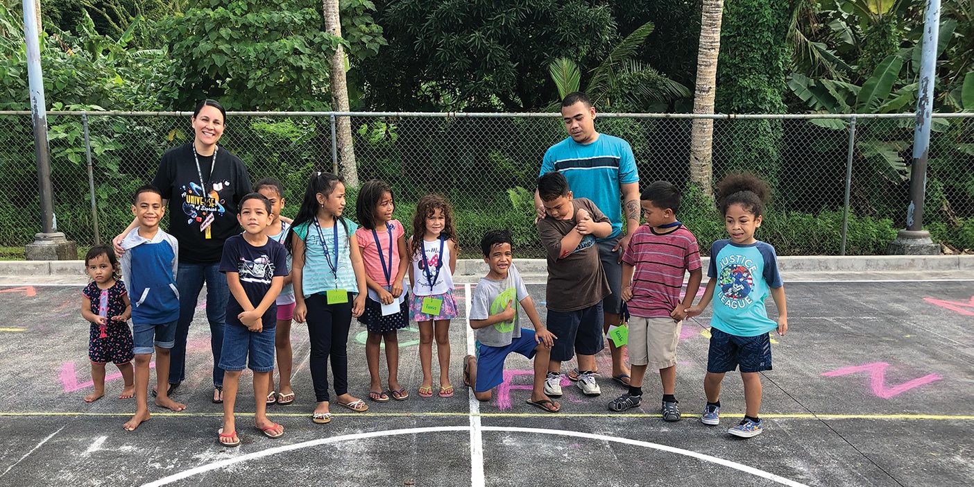 A group of American Samoan kids and volunteers stand in a line on a cement basketball court.
