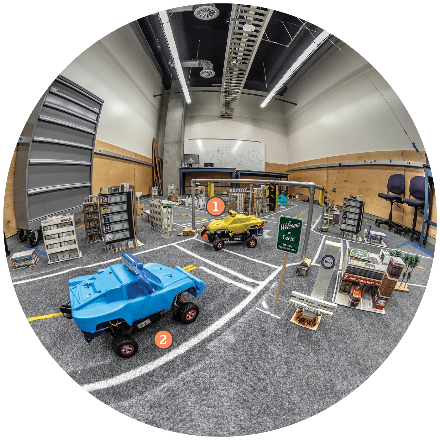 Electrical and computer engineering professor D. J. Lee's self-driving car lab, shot with a fisheye camera lens. A miniature city show two bright colored cars. The camera atop the yellow car has a number 1 annotation near it. A second blue car has a number 2 annotation near it.