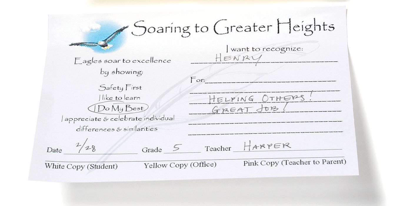 A white note written by a teacher to praise a student's good behavior. It says, "Helping others! Great job!"