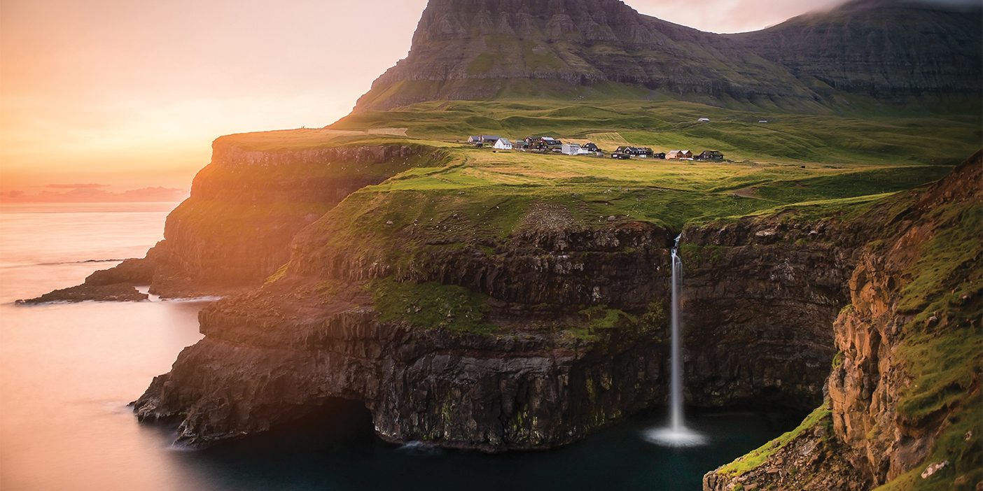 The Múlafossur Waterfall in the Faroe Islands. The sky is pink and orange, with the glow of dusk illuminating parts of the cliff's edge. On the right, a few houses sit atop a green field of grass, and then at the bottom half edge of the cliff, a silky waterfall.