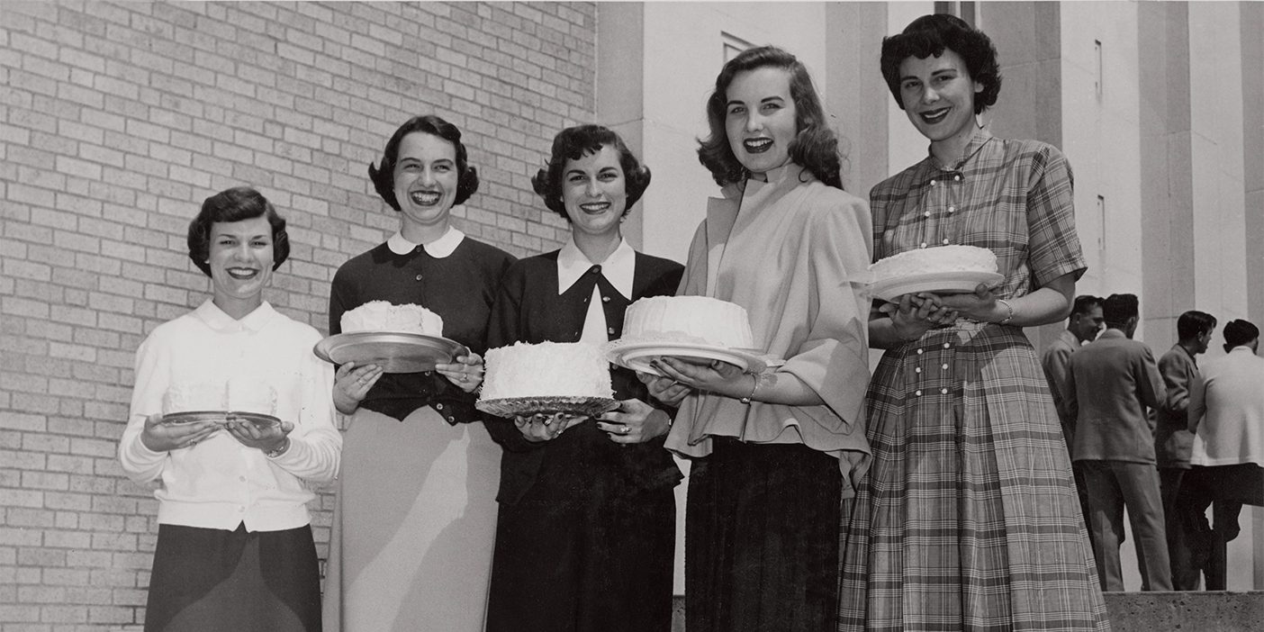 Five young women from the 1950s stand in a line, holding their cake creations, which they baked for a competition.