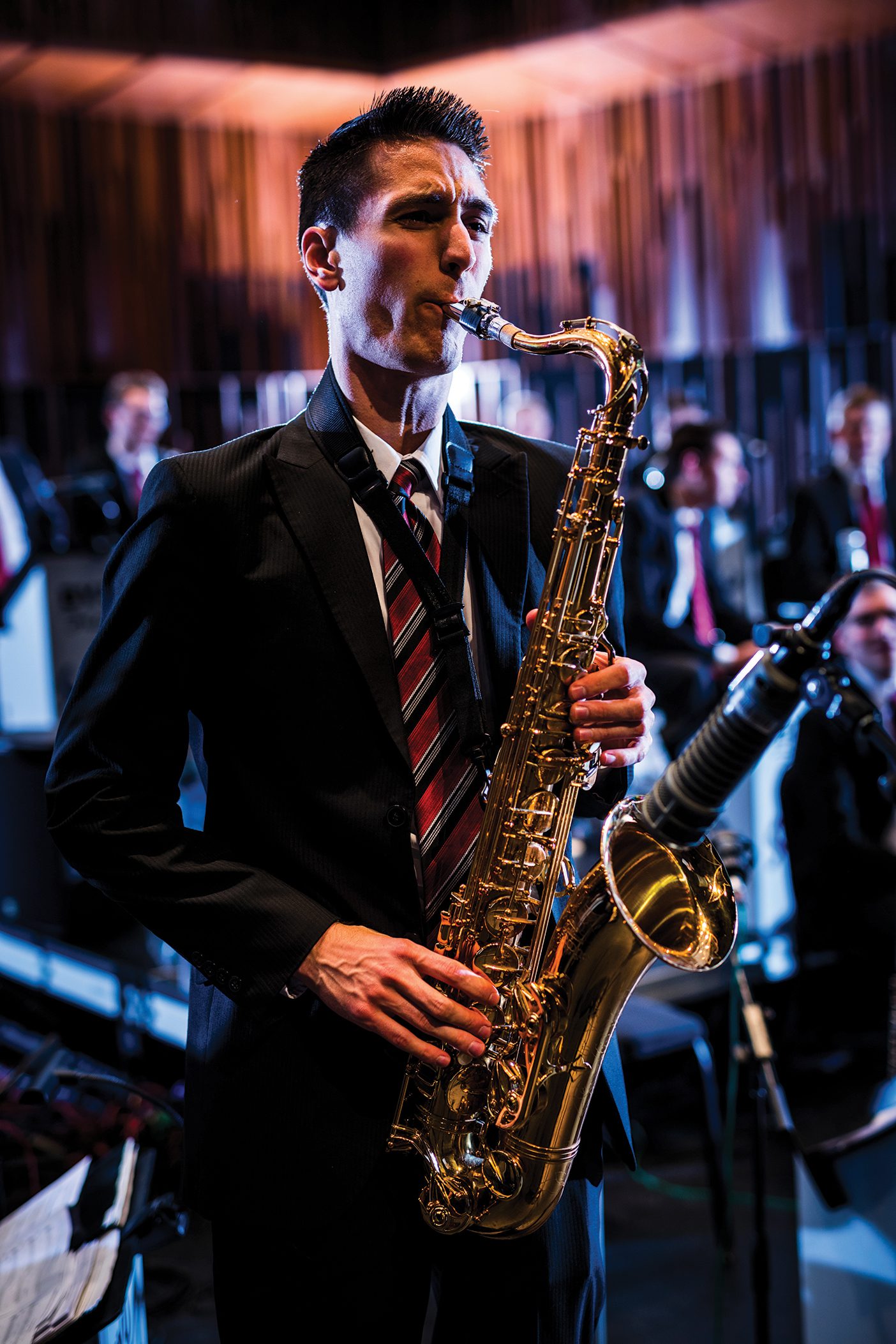 A man in a black suit with spiky hair is playing a saxophone. He's in a dimly-lit room and there are other musicians behind him dressed in black suits too.
