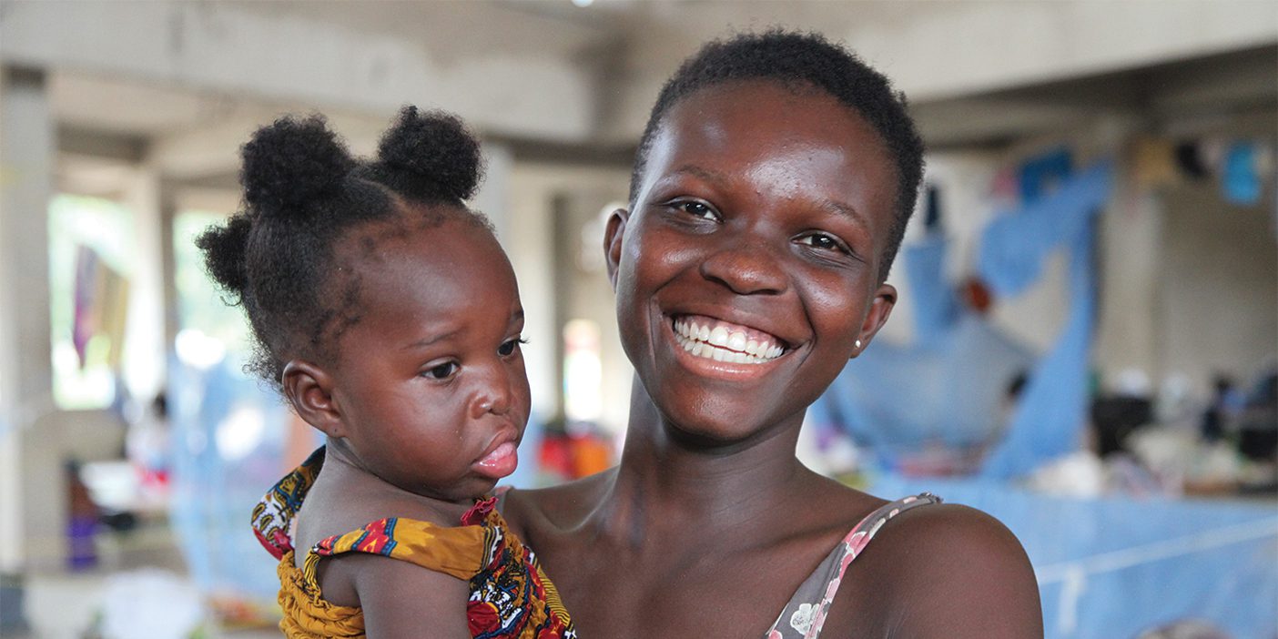 A young mother holds her toddler and smiles at the camera. The child had reconstructive surgery for a cleft palate.