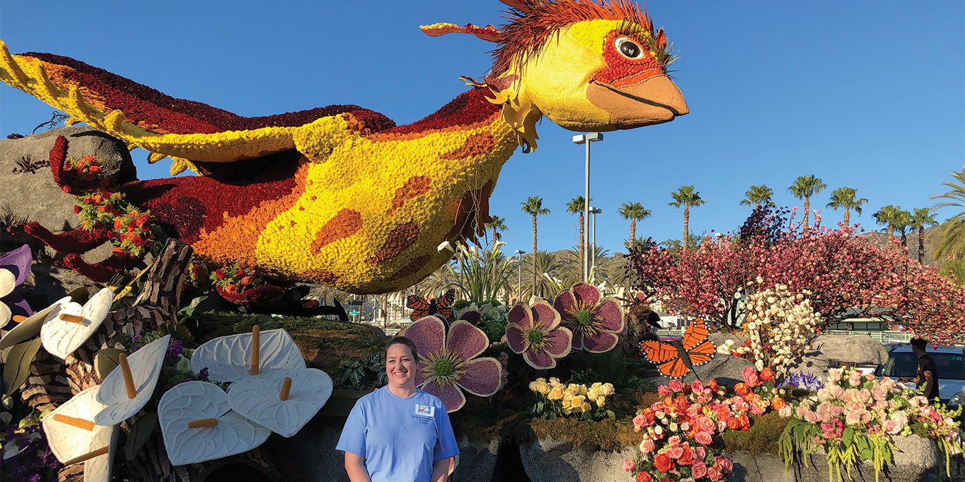 A woman in a light blue t-shirt and black pants stands in front of a parade float she created. The float is a creation of nature, with green grass, gray stones, yellow, pink, and purple flowers. Sitting atop the float is a yellow and orange bird with wings spread wide.