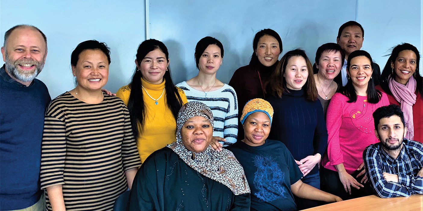 A group of 13 people from all cultural backgrounds sit and stand in front of a light wooden table. The man on the far left dressed in a blue sweater and khaki pants is Ned Gardner, who teaches English and life skills classes to immigrants in NYC.