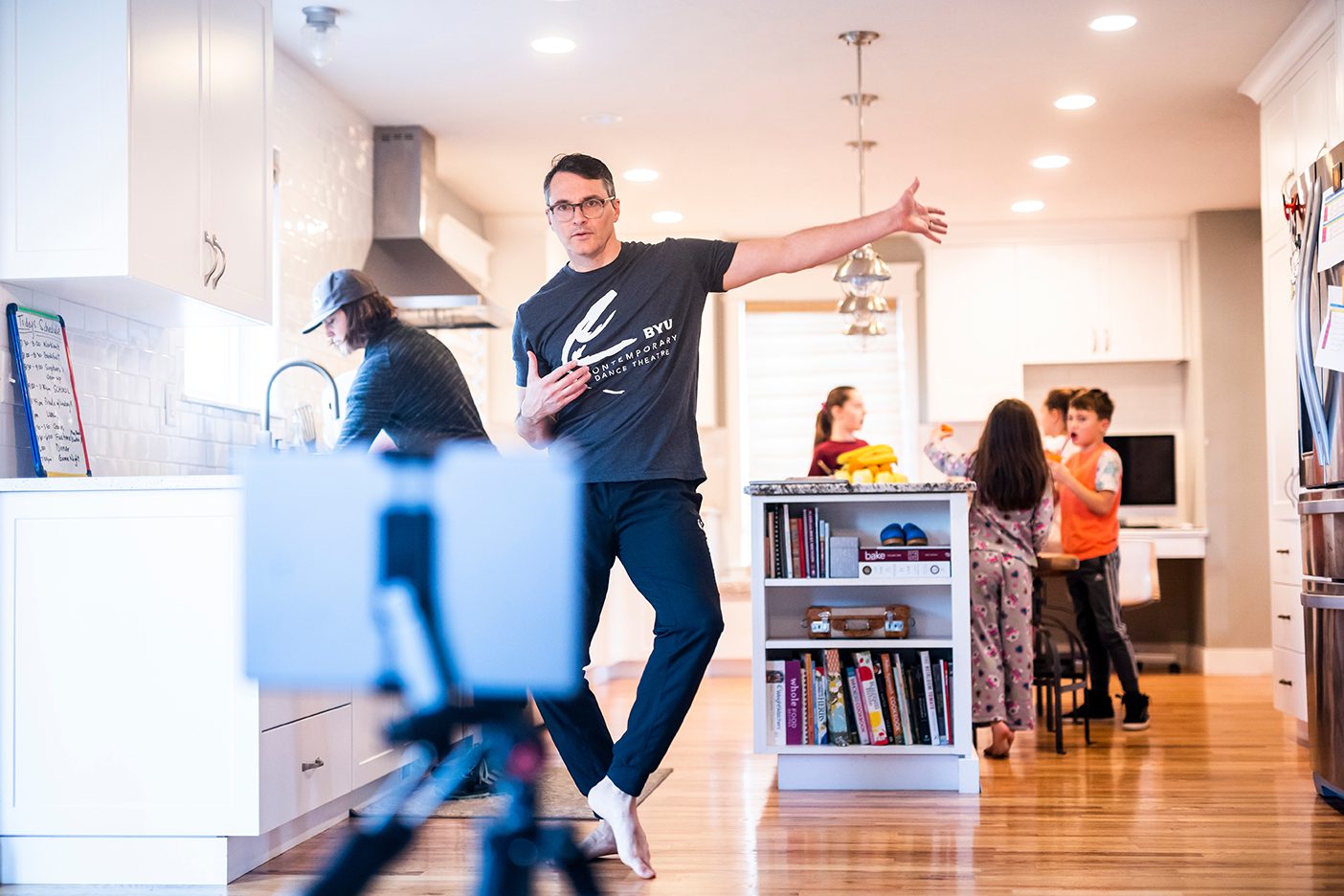Dance professor Nathan Balser dances in front of a camera in his kitchen with his kids behind him.