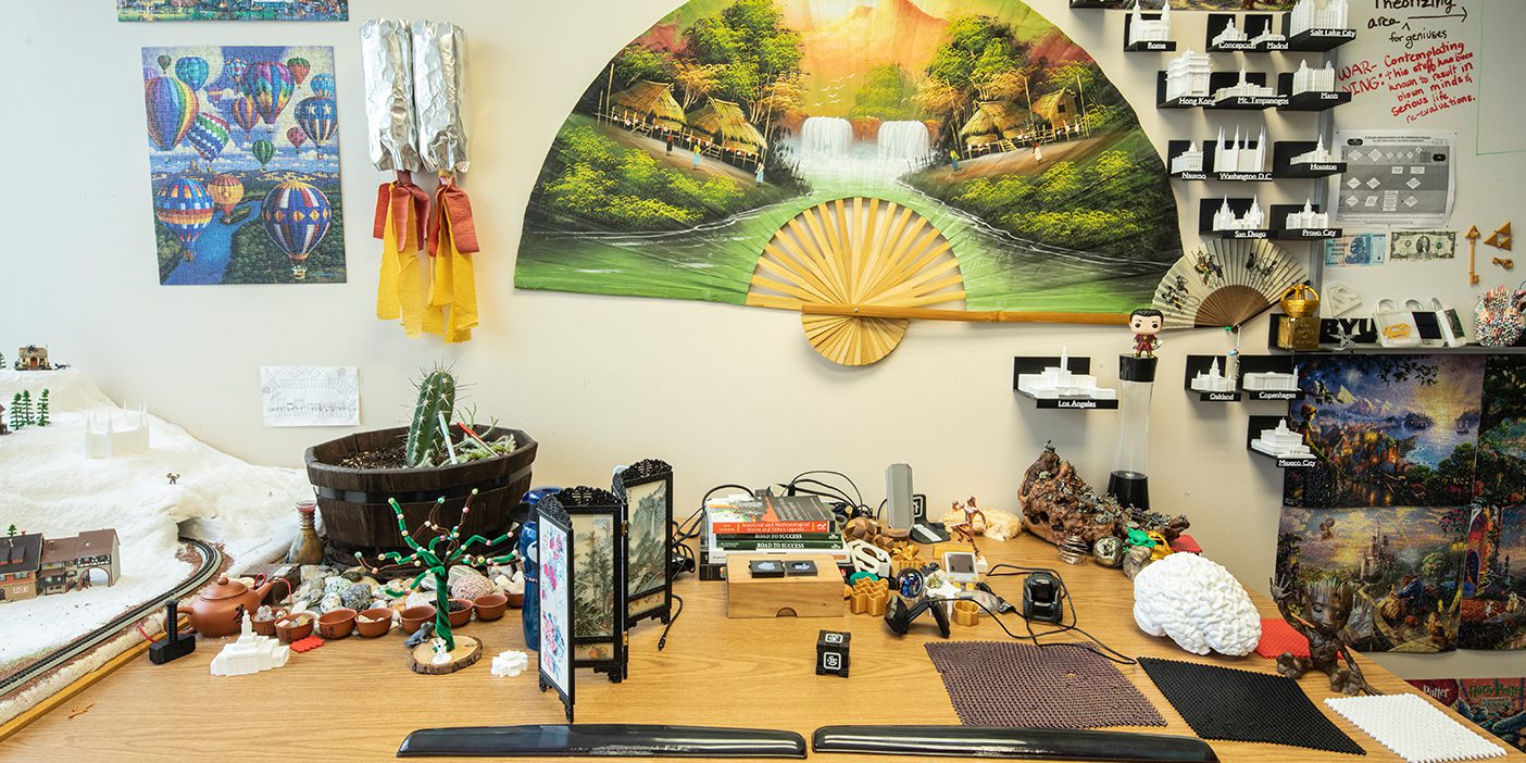 James Gaskin's desk full of rocks, plants, and other knick knacks. There is a large oriental flag on the wall and a dozen 3D-printed temples.