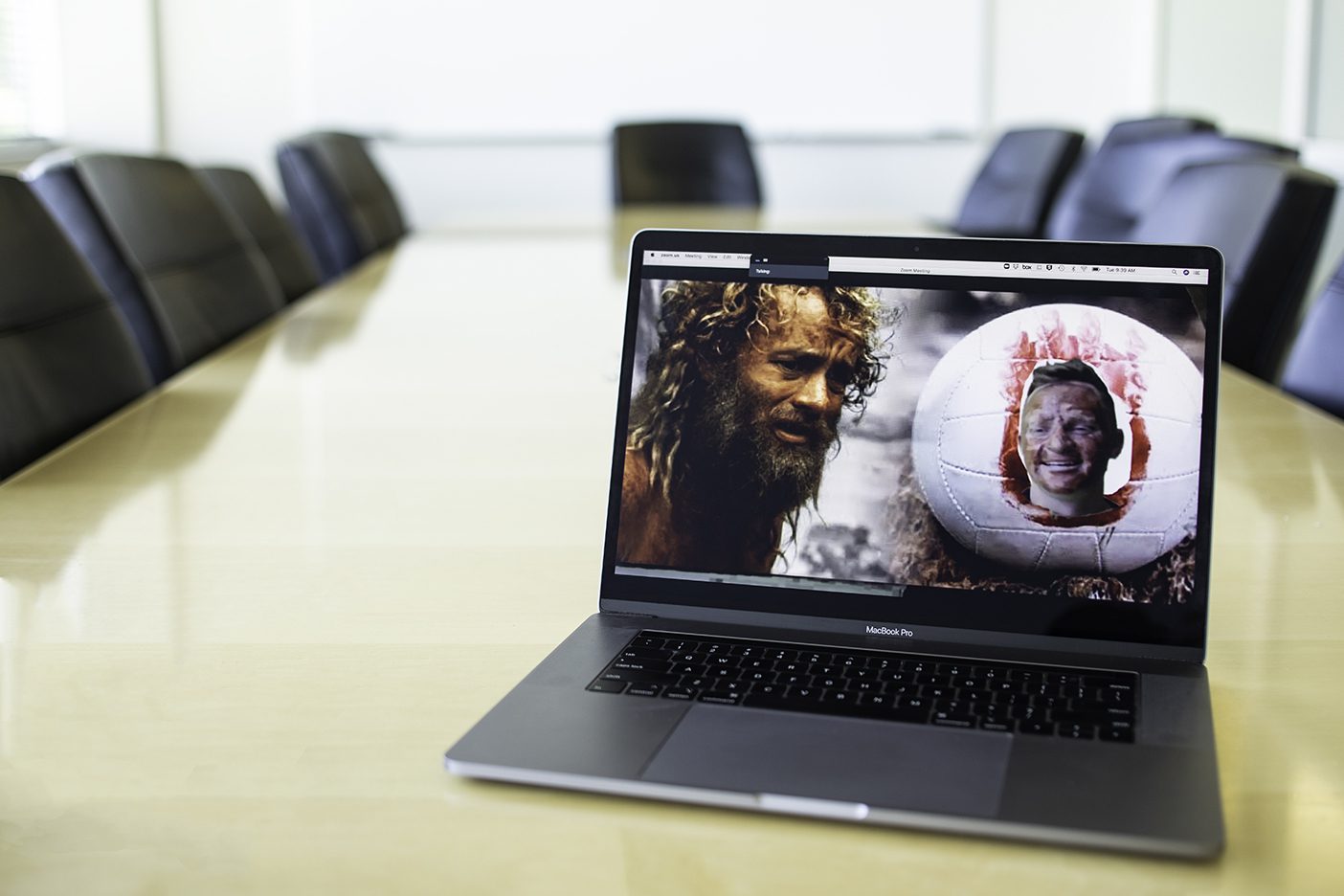 A laptop in an empty conference room with a Zoom screen showing BYU MBA student Trevor Lemmons dressed up as Wilson the volleyball from the movie Cast Away, with a Zoom background showing Tom Hanks's character beside him.