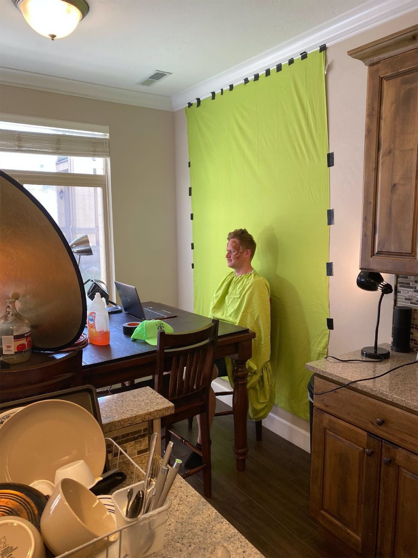 A behind-the-scenes shot of Trevor Lemmons in a green shirt and with a green screen to be Wilson the volleyball on a Zoom call.
