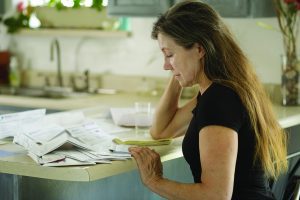 A woman sits at her kitchen counter looking thoughtfully at a stack of bills.