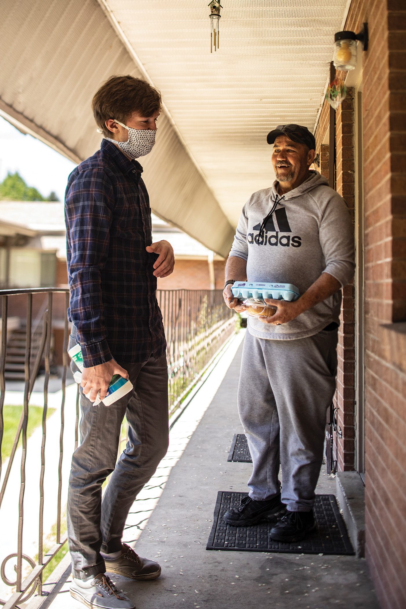 John Lindsay, wearing a protective mask and a plaid button-down shirt, is standing just outside the doorstep of a second-story apartment. He is speaking to the apartment-owner, a middle-aged man with a grey sweatshirt and black baseball cap. The middle-aged man is holding a carton of eggs and smiling.