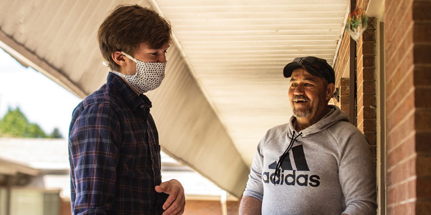 A young man wearing a protective mask and a plaid button-down shirt is standing just outside the doorstep of a second-story apartment. He is speaking to the apartment-owner, a middle-aged man with a grey sweatshirt and black baseball cap. The middle-aged man is holding a carton of eggs and smiling.