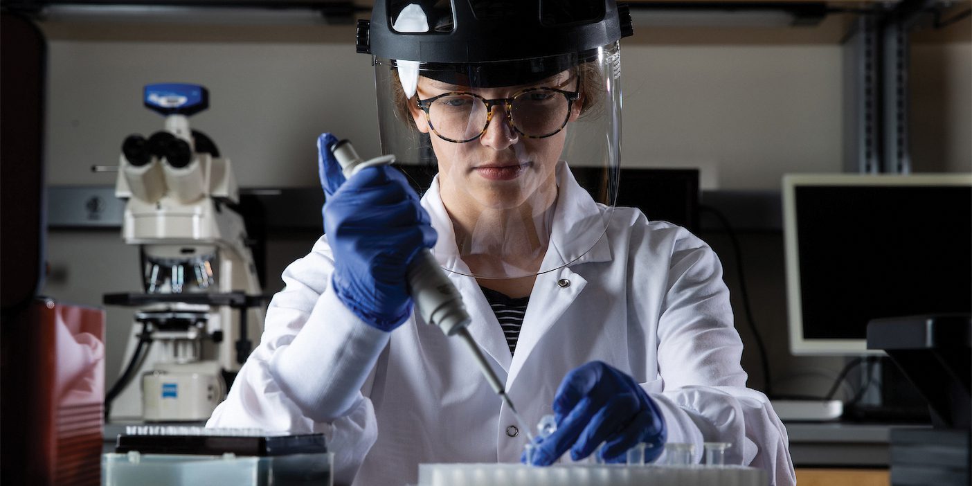 A young female scientist in protective gear uses a tool to extract a substance from a clear container.