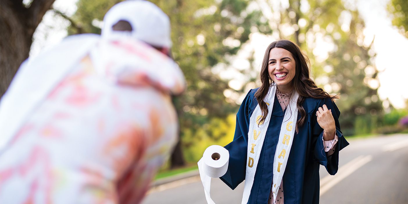 A female BYU grad smiles while wearing a graduation stole made from toilet paper with the word "COVID GRAD" on it.