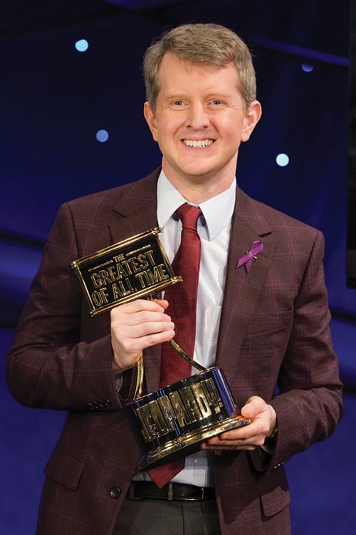 Ken Jennings III stands smiling proudly, holding the trophy declaring him the Jeopardy! GOAT.