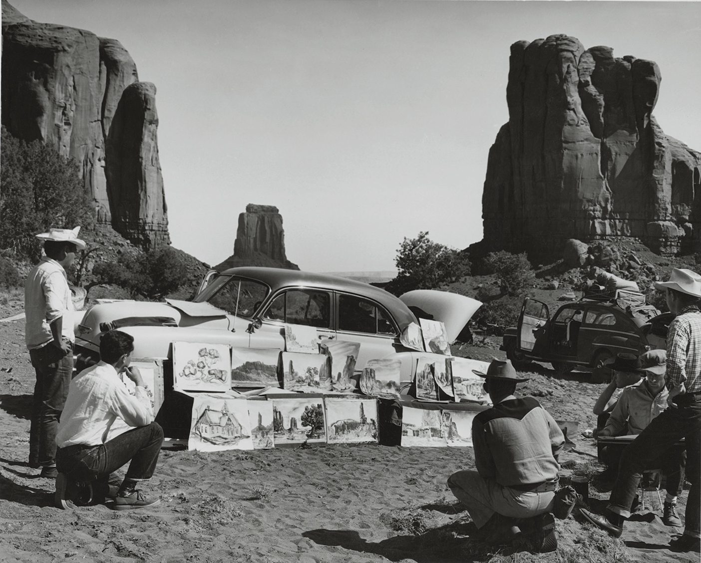 Art students look at a collection of art pieces leaning against a car at the base of a red-rock landscape.