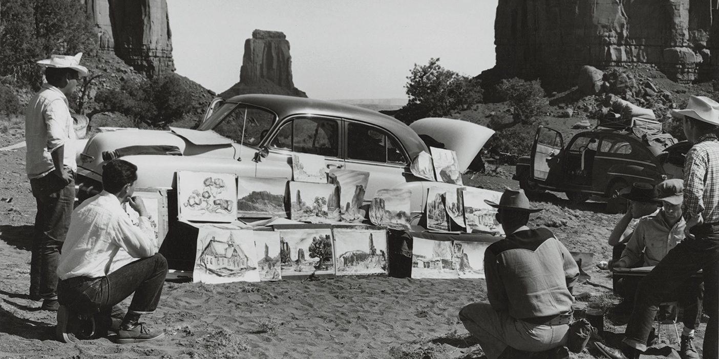 Art students look at a collection of art pieces leaning against a car at the base of a red-rock landscape.