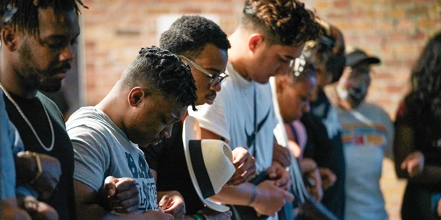 A row of people link arms and boy their heads in prayer at a event sharing the experiences of people of color.