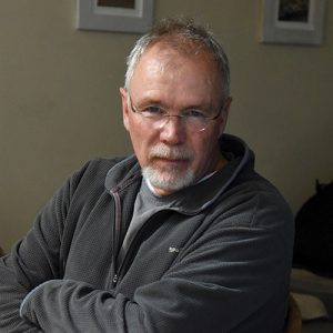 A man in his fifties with thinning gray hair and a beard wears glasses and a dark gray jacket as he looks at the camera.