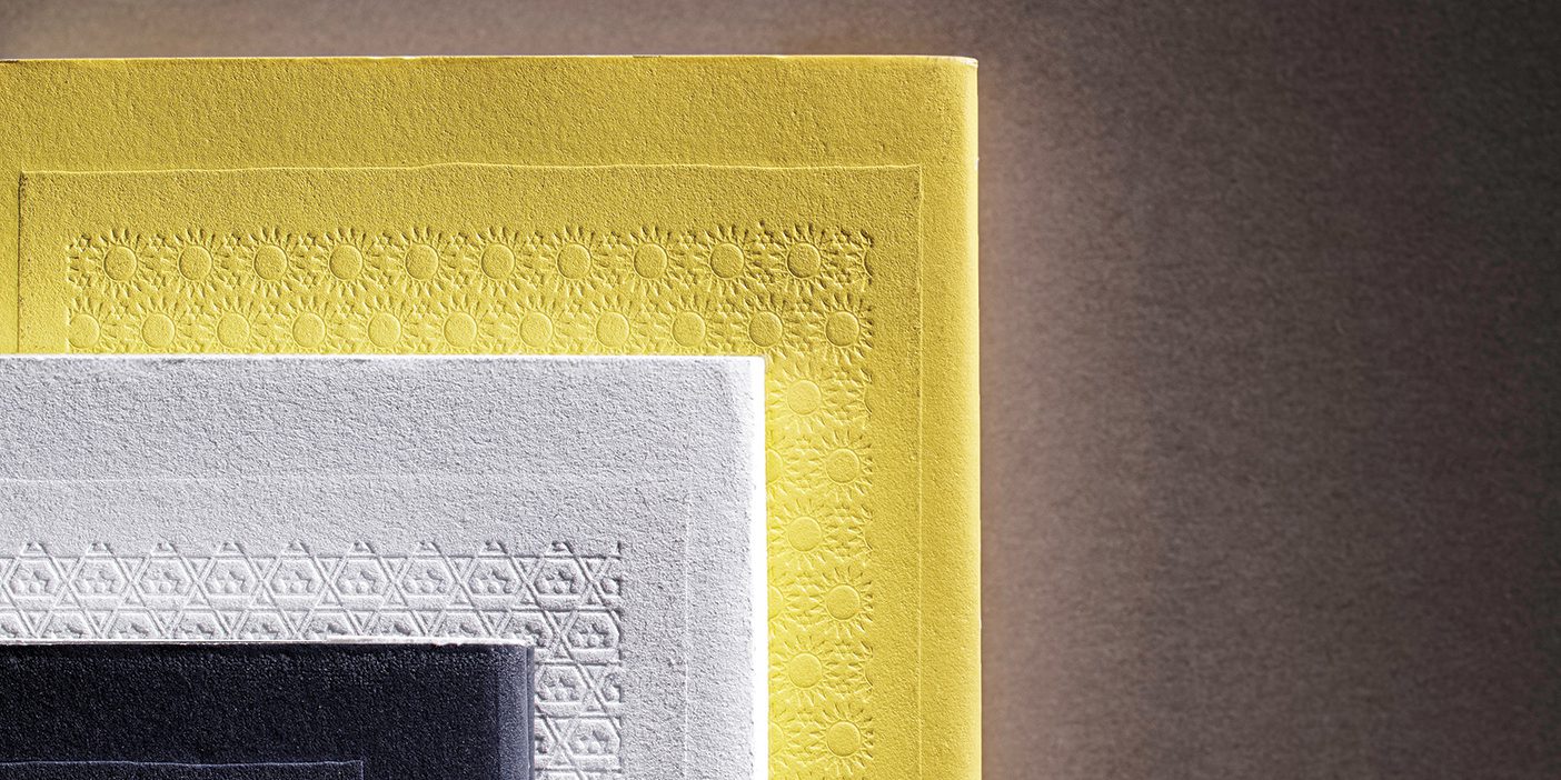 Three books are stacked on top of each other; a small black book, a medium-sized white book, and a large yellow book. The covers are embossed with intricate designs.