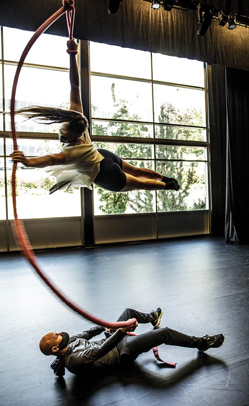 Adam Dyer lays on the floor, holding a long rope that twirls dancer Mariah Sainsbury parallel to the floor.