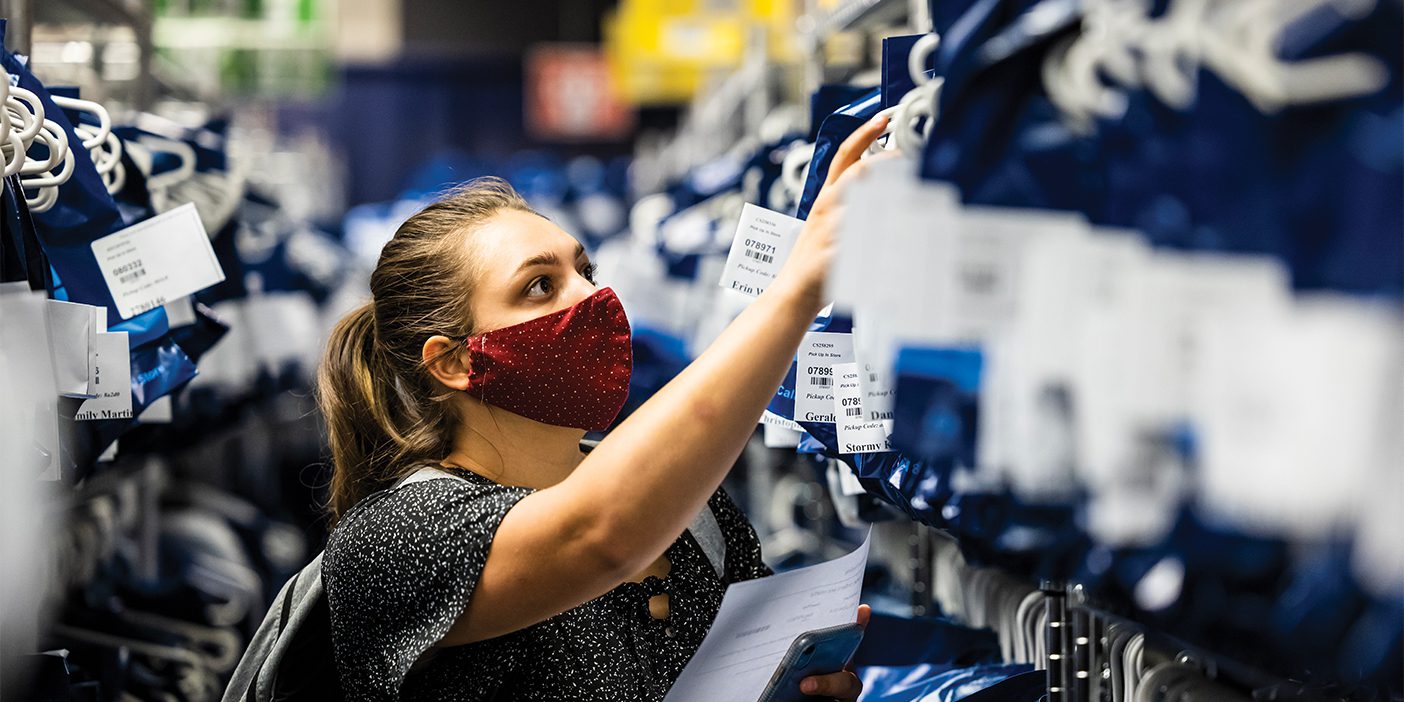 A female student wearing a cloth face mask sorts through a row of blue bags containing textbooks.