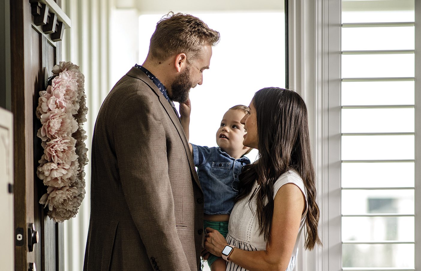 A mom and child say goodbye to a man dressed in a sportcoat, sweatpants, and bare feet at the front door of their home.