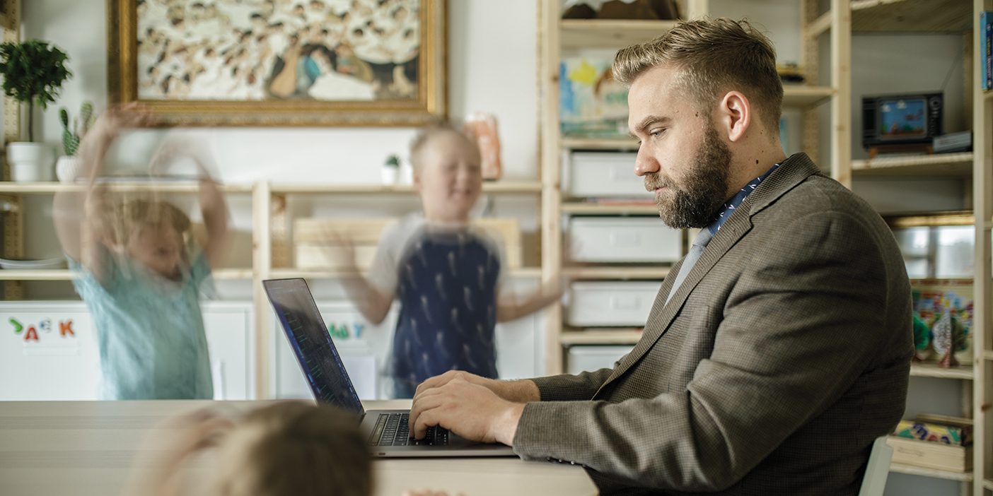 A man concentrates on his laptop at a table flanked by three children who are trying mightily to distract him.