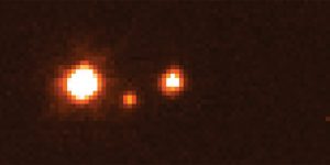 A pixelated image with an orange filter featuring Haumea and its moons. Haumea is a bright white/orange pixelated blob on the left. Its moons are smaller orange pixels on the right.
