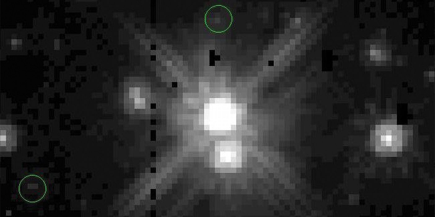 A black and white pixelated image featuring Pluto and its moons. Pluto is a bright pixelated white dot in the center. The moons are dim grey pixels on the outskirts of the image. The moons are circled in green.