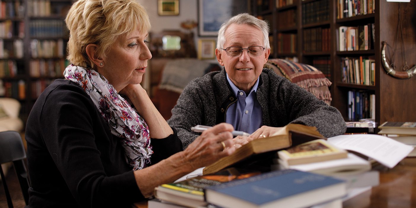 Fiona and Terryl Givens sit side by side at a table in their home library. Their table is covered in books. Fiona holds a pen in hand, about to mark a passage from a book.