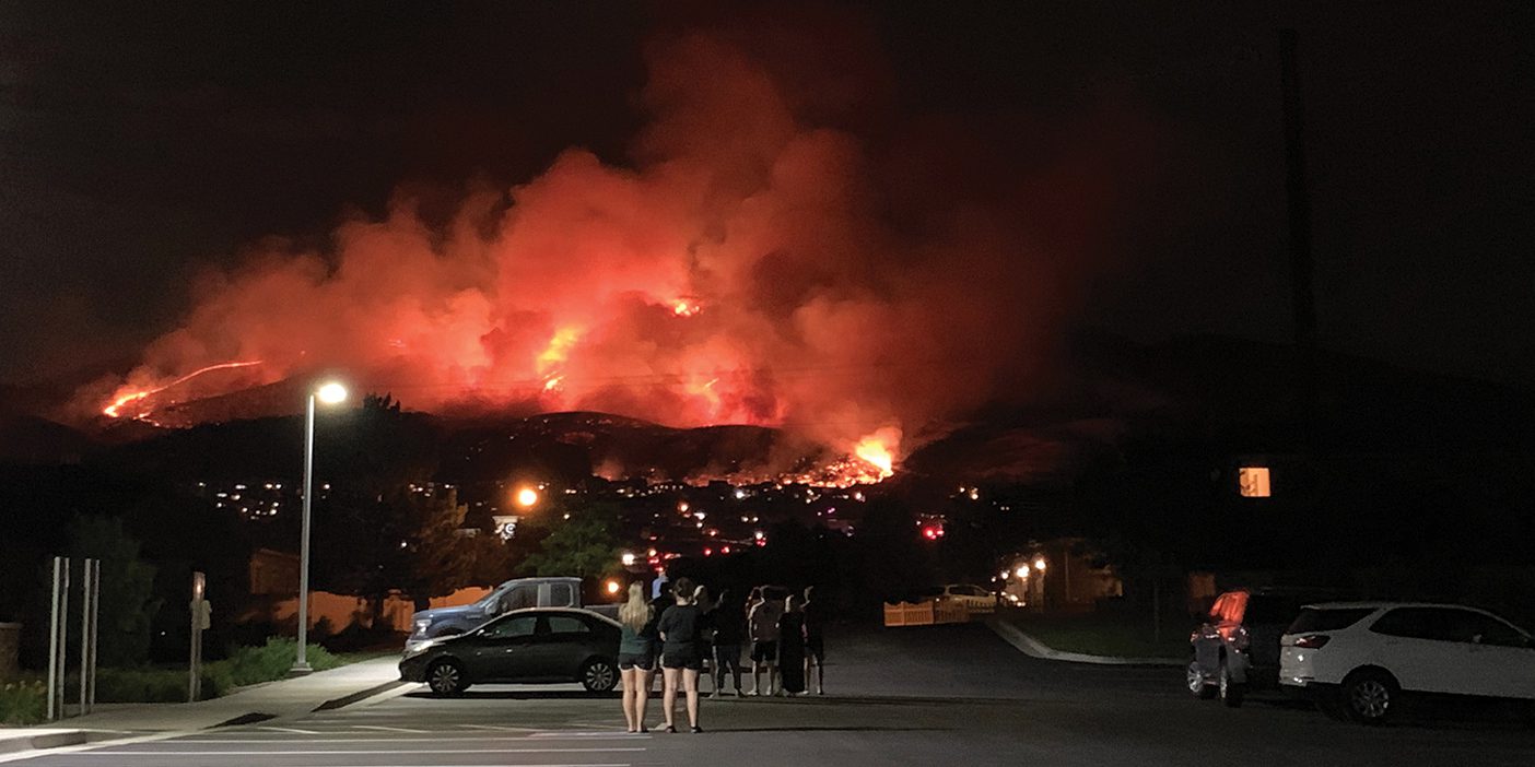 People stand in a parking lot at night, looking at a mountainside glowing with orange fire and smoke.