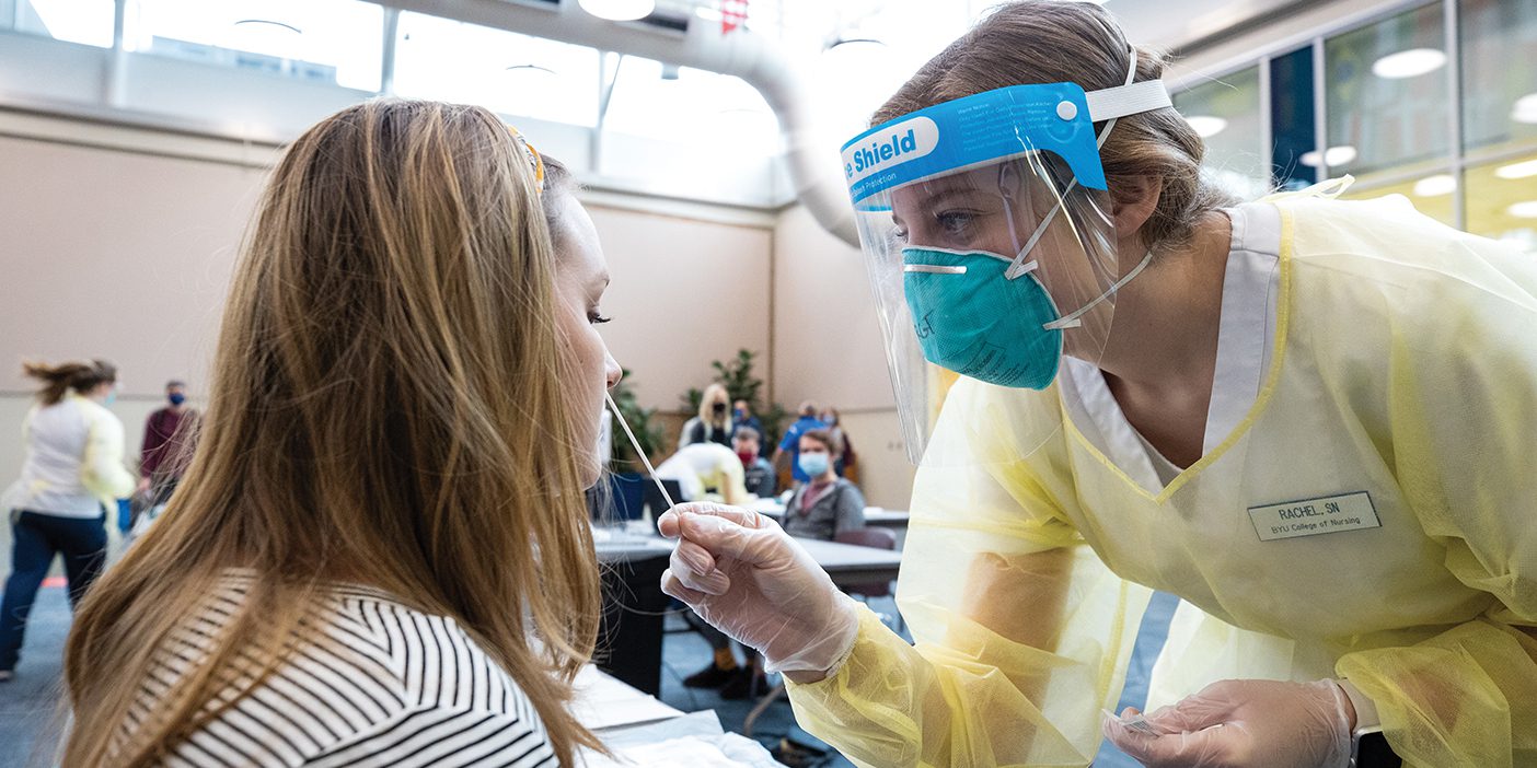 A female nursing student wearing mask, gloves, gown, and face shield inserts a swab into the nostril of a female student to test for COVID-19.