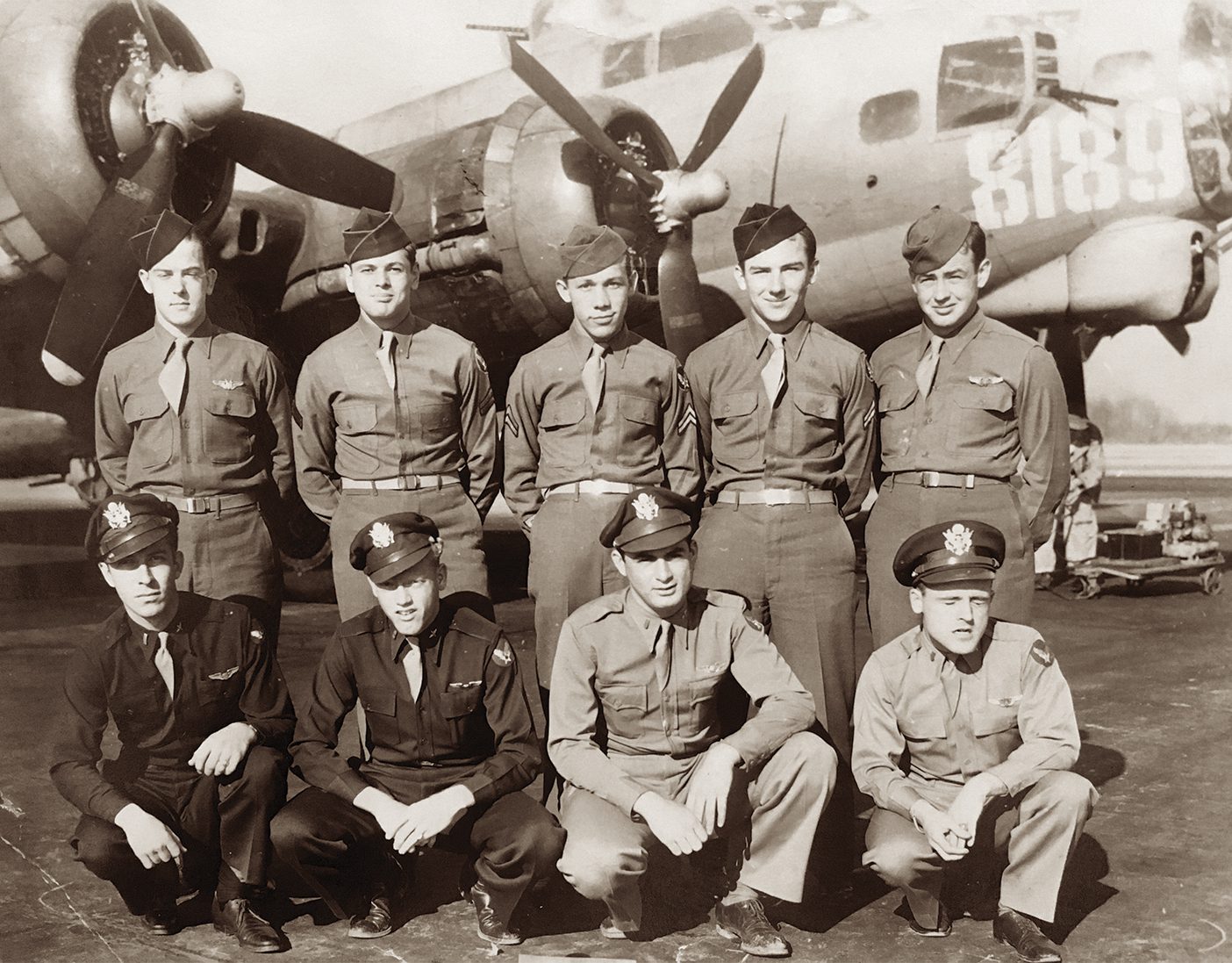 A sepia photo of nine men in military uniforms posing in front of a WWII airplane.