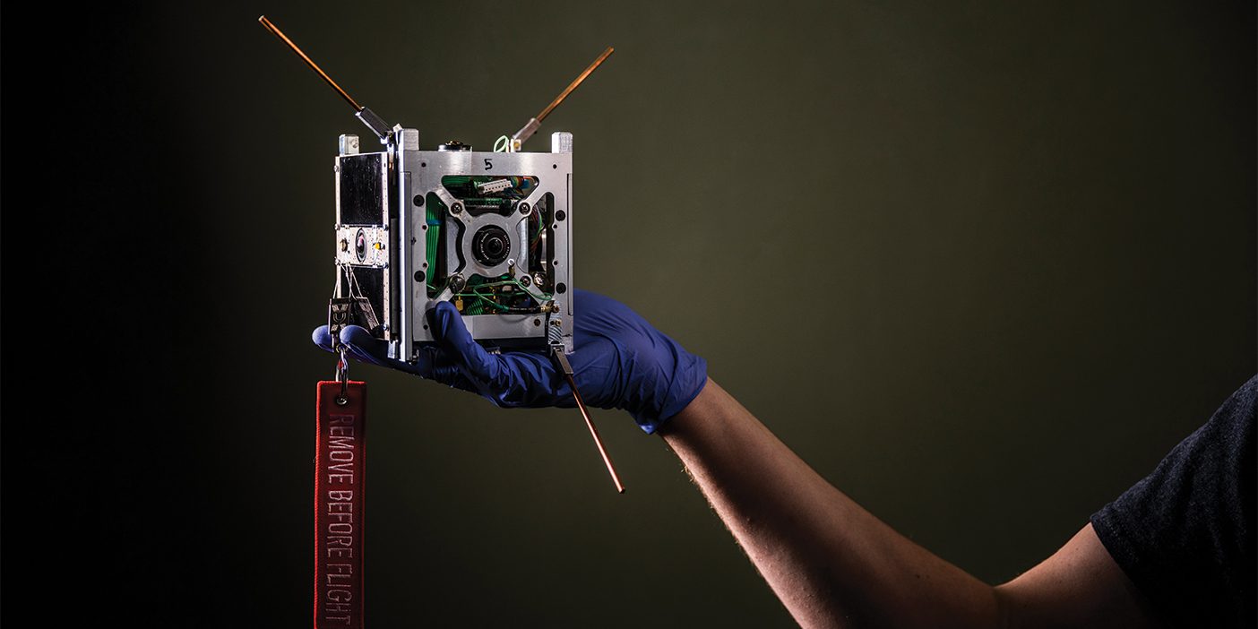 A CubeSat—a small satellite that looks like a metal cube—being held by a hand.