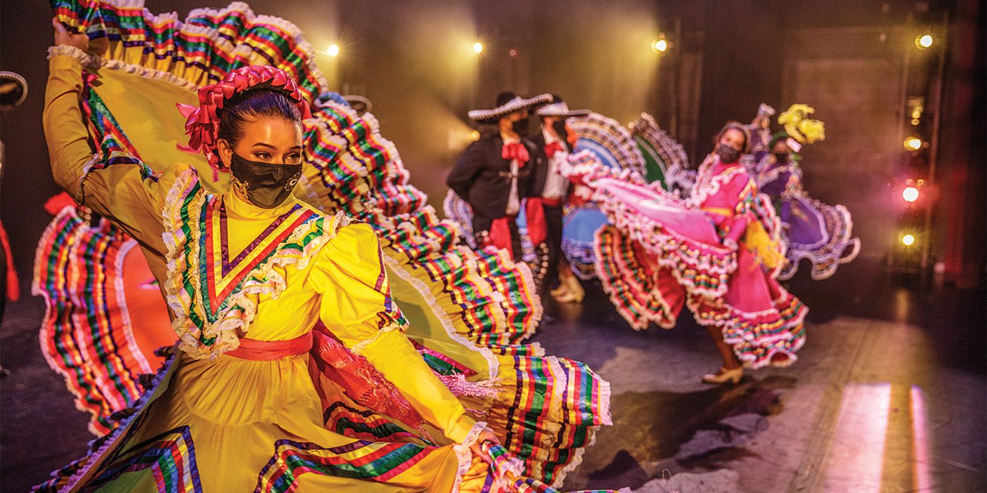 Male and female dancers wearing colorful traditional Central American costumes perform on stage.