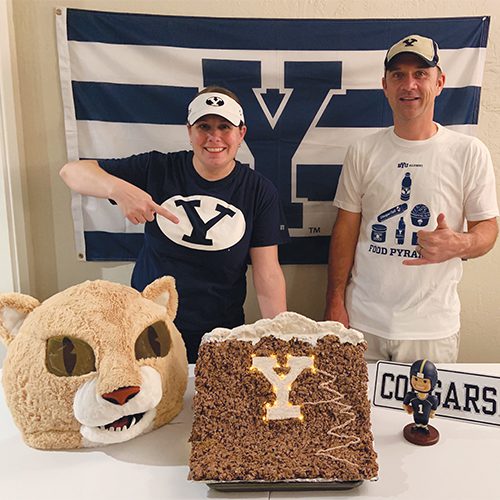 Two BYU fans stand in front of a BYU flag. They point to the table in front of them, which displays a Y Mountain replica made from rice crispy treats. The Y on the mountain is illuminated.