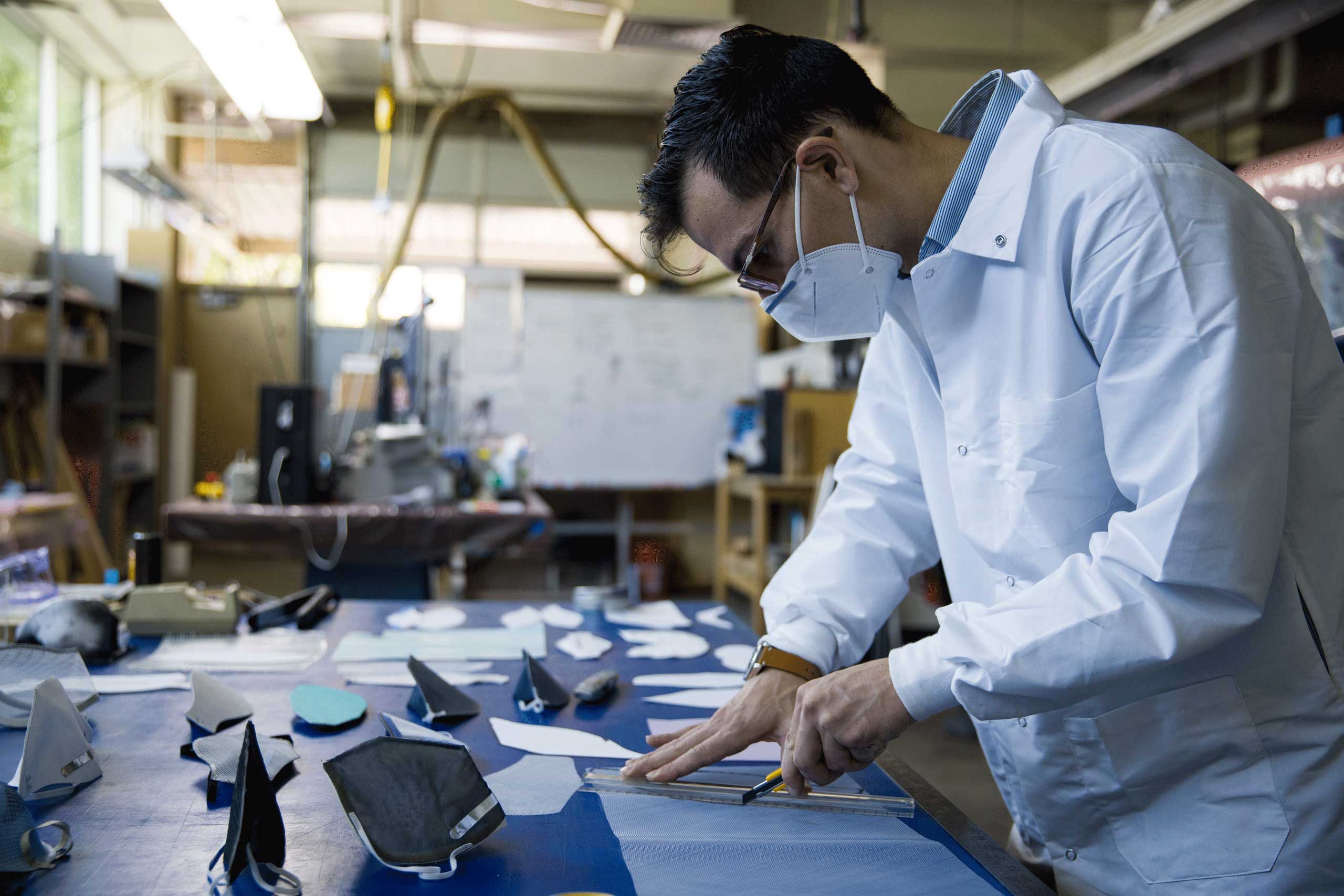 A male student in a white lab coat, wearing a mask, cuts fabric with a small blade to make cloth masks.