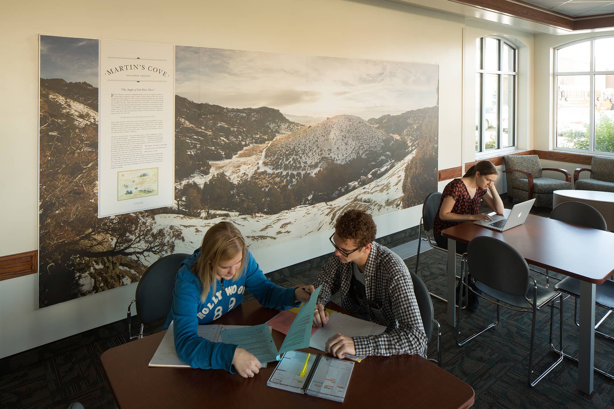 Two students sit at a table studying in front of a mural of Martin's Cove in BYU's Heritage Halls.