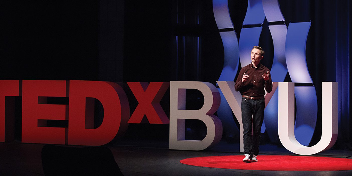 Dave Vance stands on the red TEDxBYU stage, gesturing emphatically to his audience.