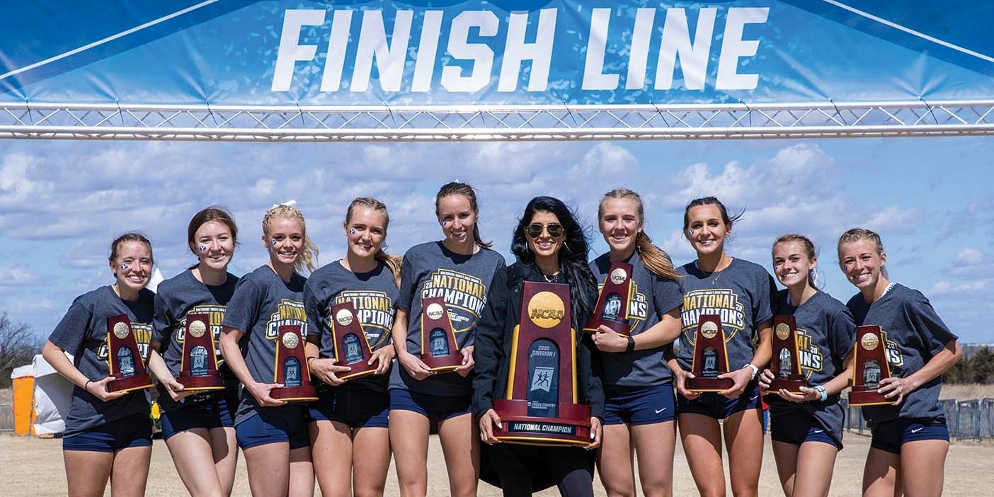 BYU Women's Cross Country team of nine stands under the finish line banner at the NCAA Women's Cross Country Championships, with Coach Diljeet Taylor in the middle, holding their first-place trophy.