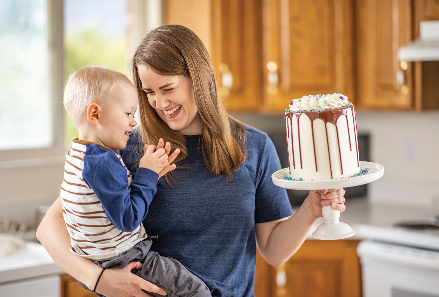 A young mom holds her toddler son in one arm and a beautifully decorated cake in the other.