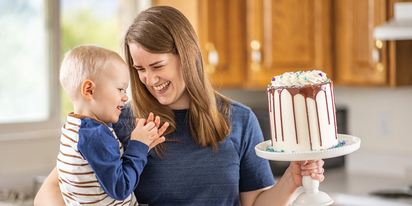 A young mom holds her toddler son in one arm and a beautifully decorated cake in the other.