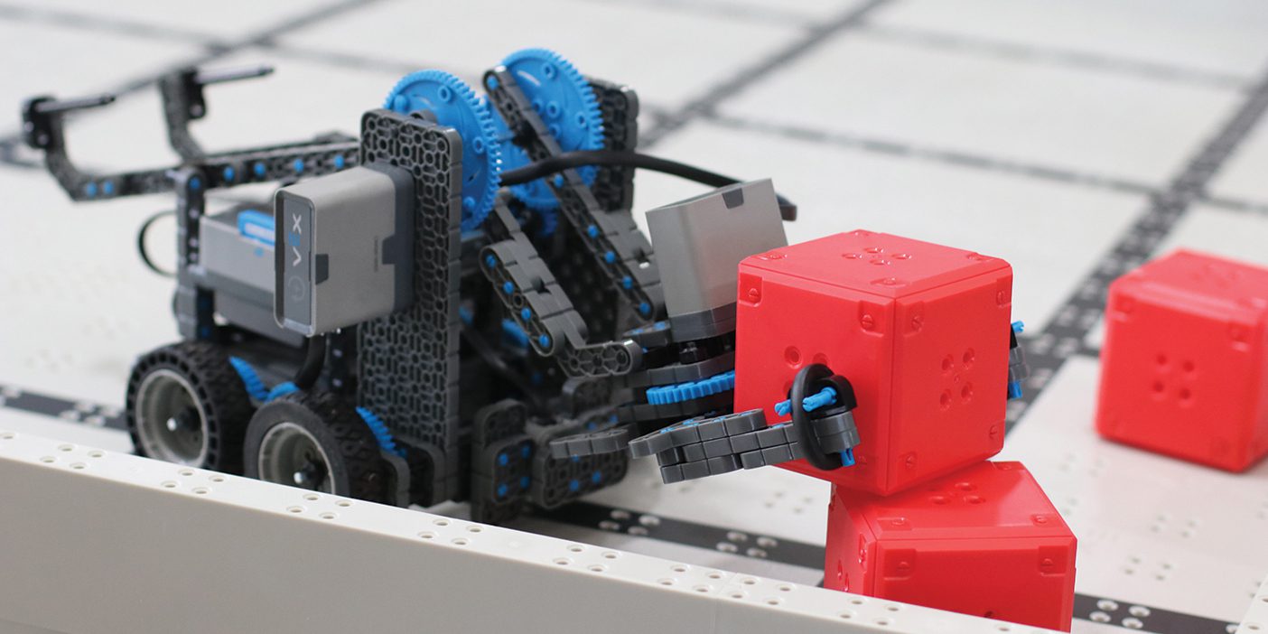 A tiny robot made of plastic parts picks up a small red block.