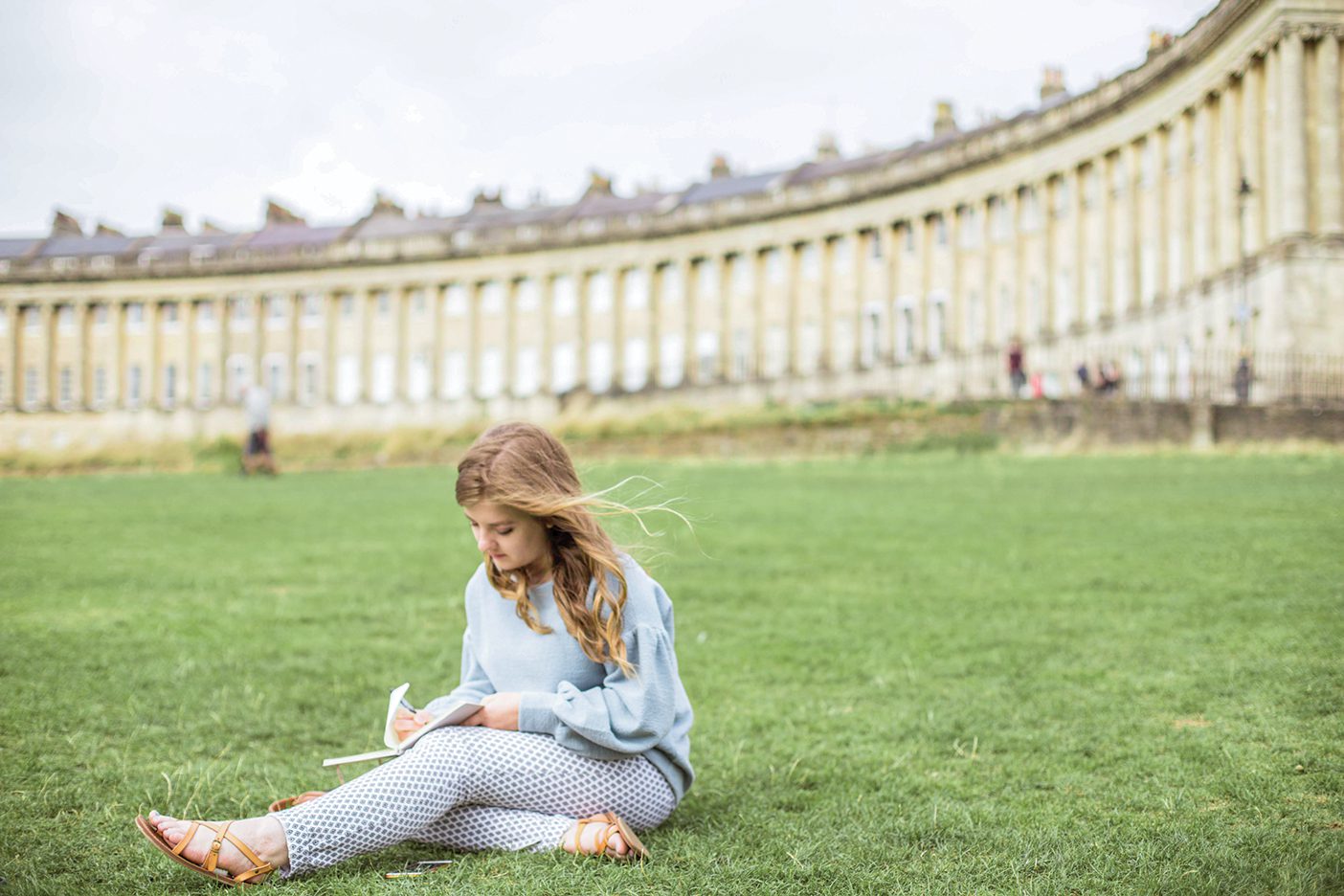A picture of a girl writing as she sits on the grass in front of the Royal Crescent in Bath.