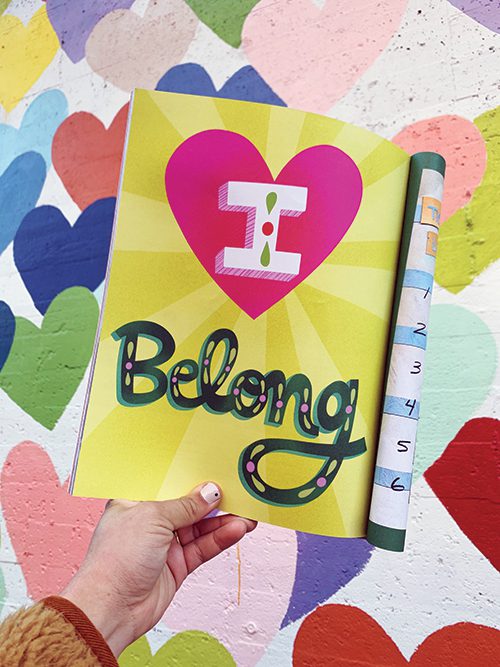 A page from Ditto Kids magazine being held in front of a wall with colorful hearts. The page says, "I Belong."
