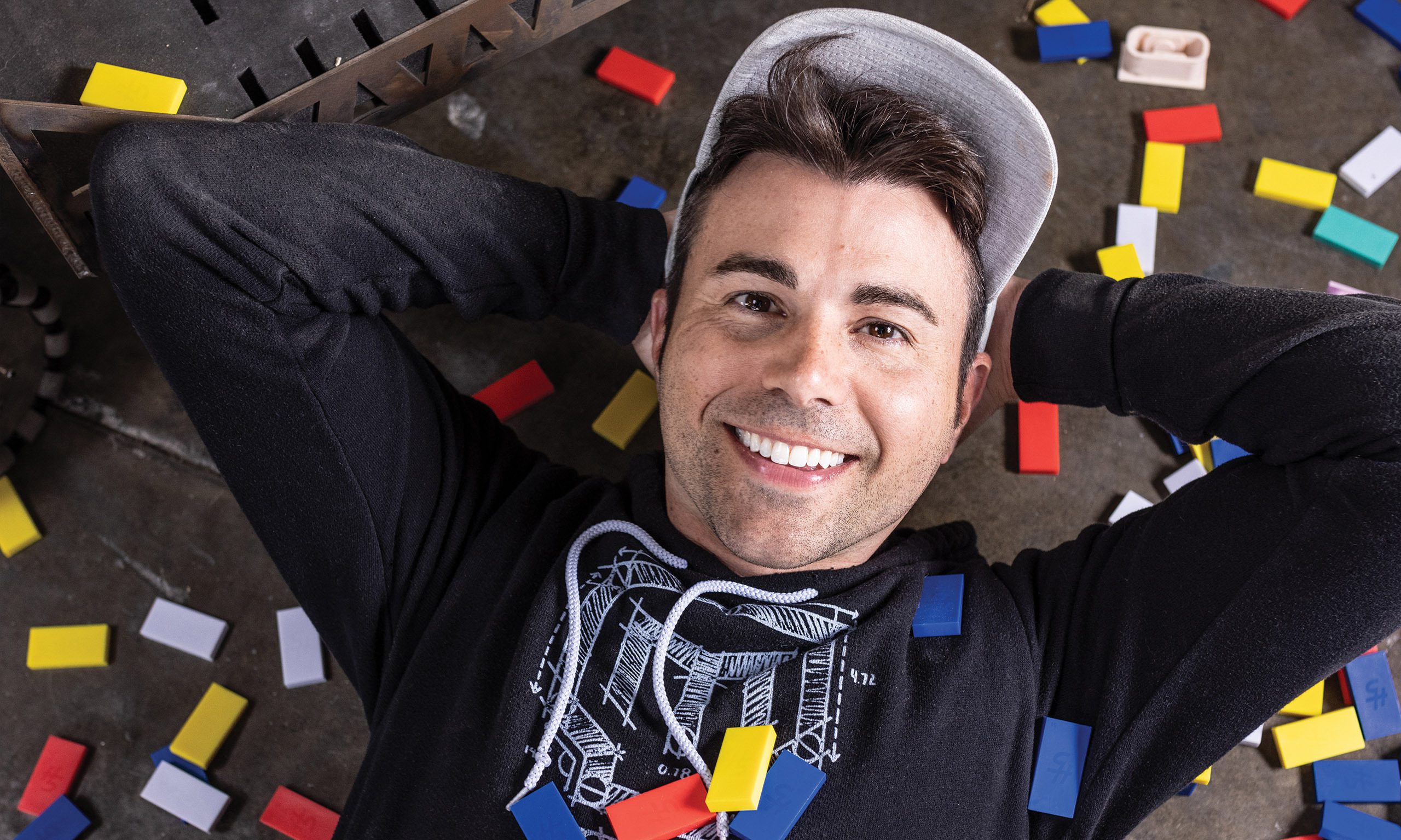 Youtuber Mark Rober laying down on a floor full of yellow, red, white, and blue dominos.