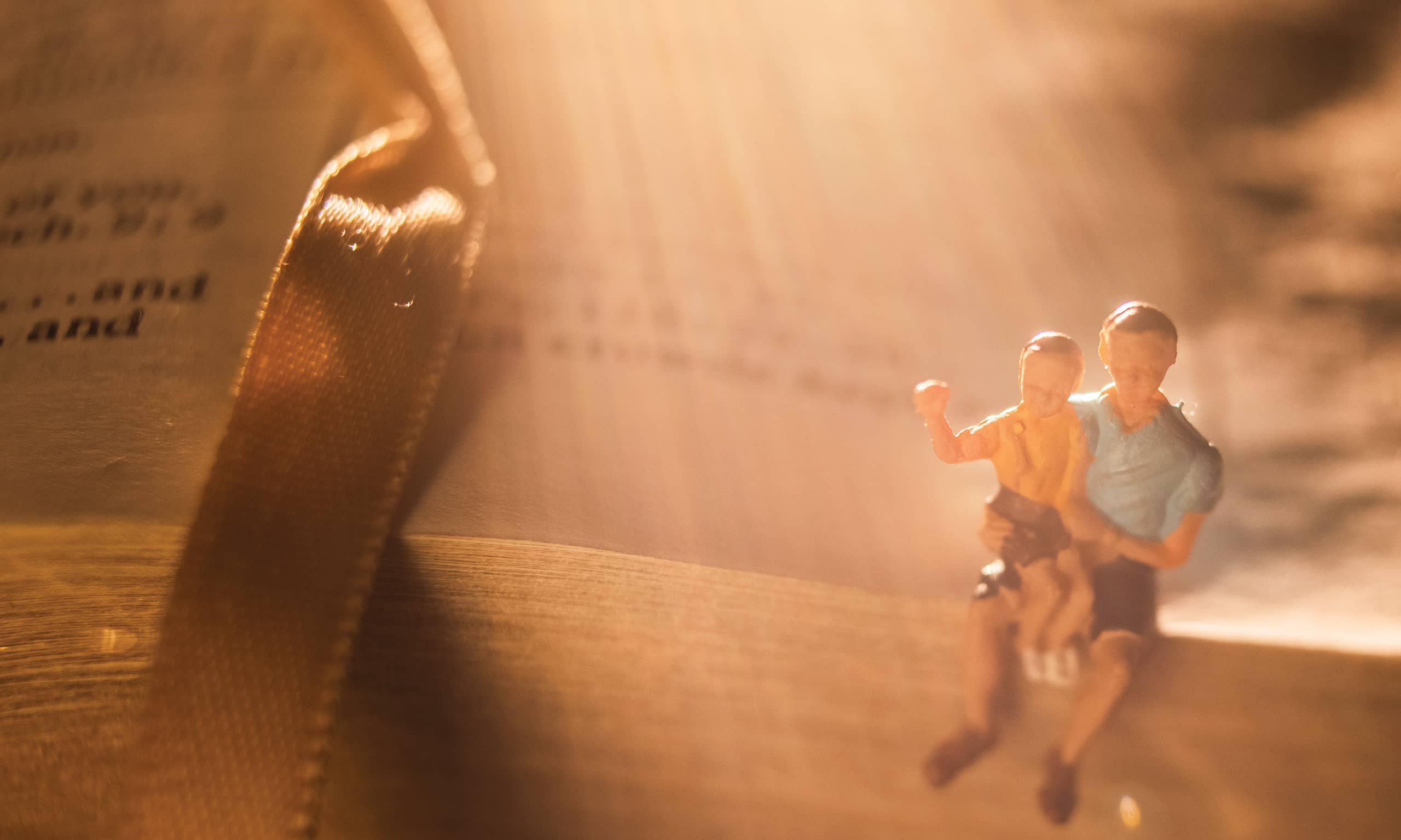 Small figurines of a parent and child sit on the edge of scripture pages bathed in golden light.