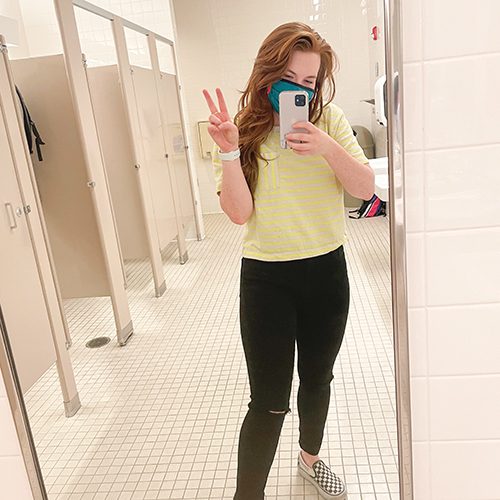 A woman stands in front of a public bathroom mirror taking a selfie. There are several stalls behind her.