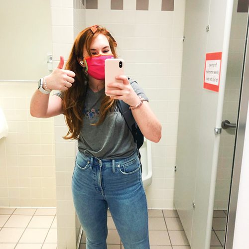 A woman stands in front of a public bathroom mirror taking a selfie.